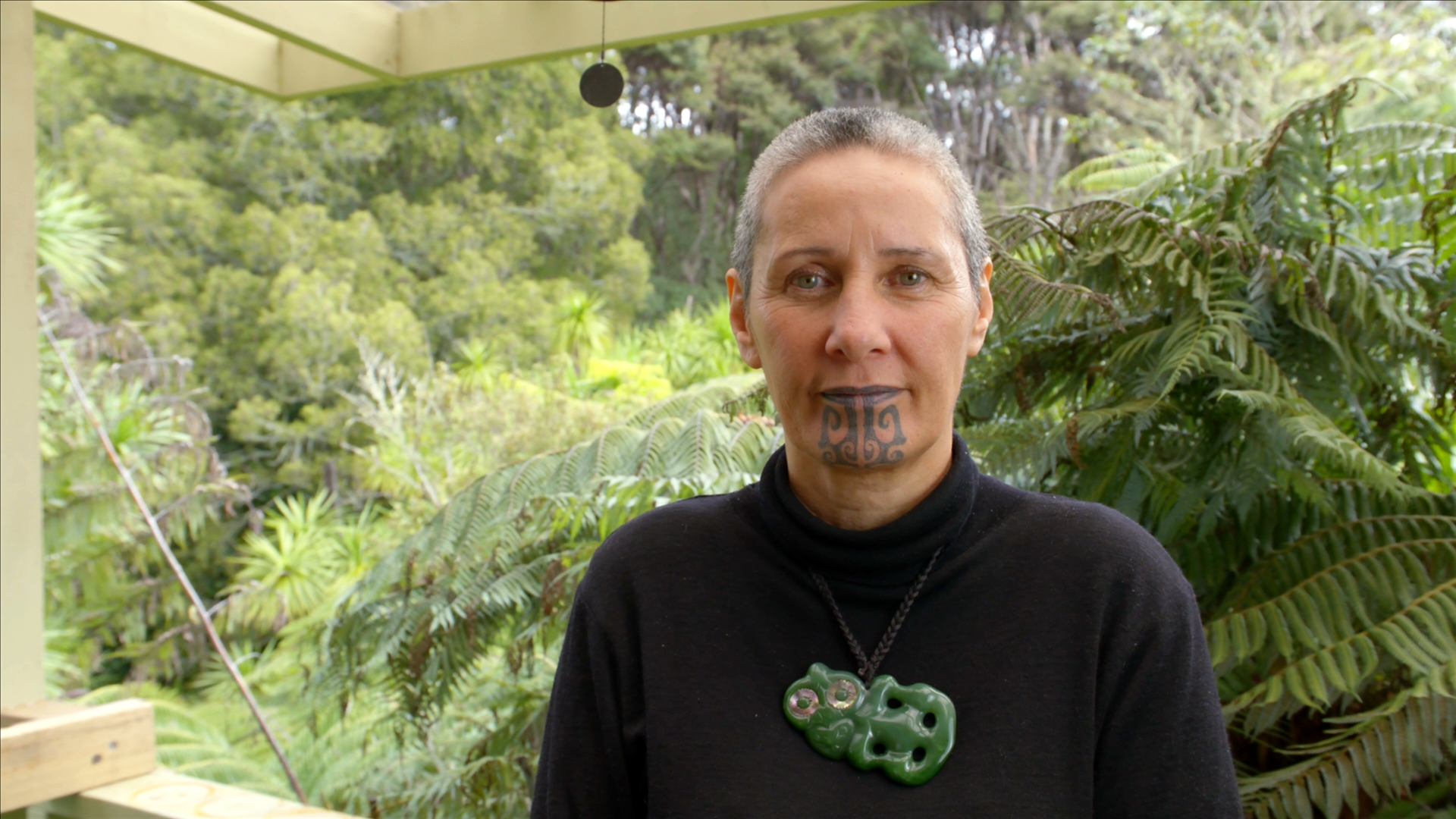 Headshot of Kehe Pene from the shoulders up, standing on a wooden structure outside in front of a dense, green garden